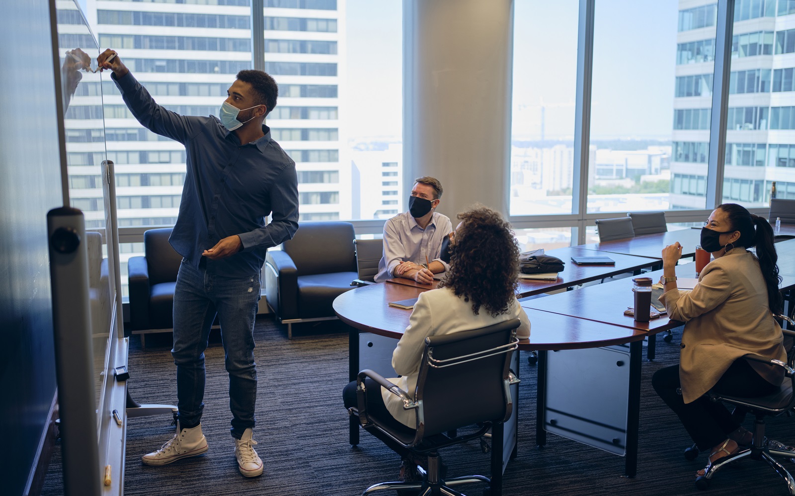 A group of people seated in an office meeting room, who are wearing face masks and in a discussion while one male stands at a white board.
